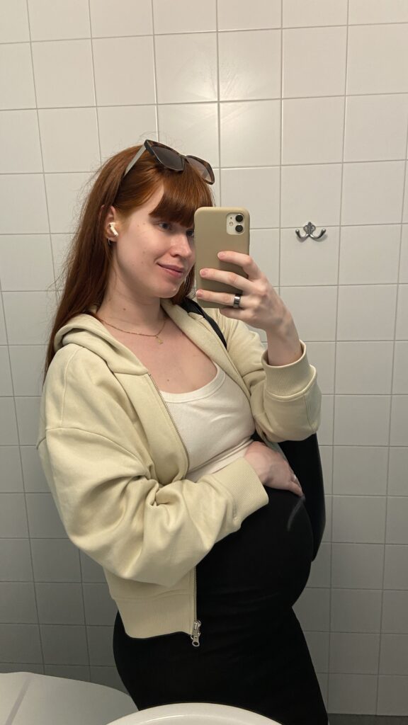 Pregnant woman with red hair taking a mirror selfie in a light beige sweatshirt from &Otherstories. 